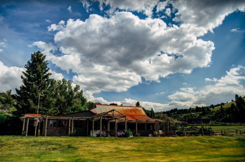 A rustic house with a red roof surrounded by green fields and trees under a blue sky with fluffy clouds.