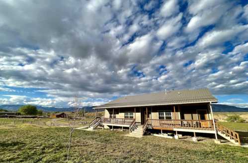 A rustic wooden house with a porch, surrounded by grassy fields and a dramatic sky filled with clouds.