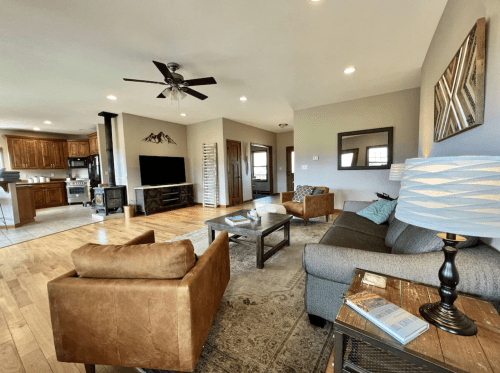 Cozy living room with a gray sofa, brown chairs, wooden floors, and a view of an open kitchen.
