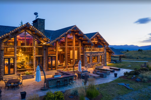 A beautifully lit log cabin-style lodge at dusk, surrounded by outdoor seating and scenic mountains in the background.