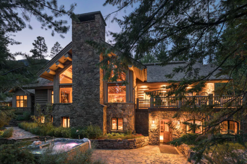 A stone house with large windows and a chimney, surrounded by trees, illuminated at dusk.
