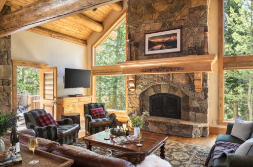Cozy living room with stone fireplace, wooden beams, and large windows overlooking trees, featuring comfortable seating.