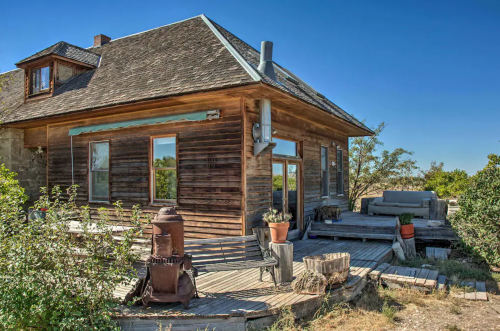 A rustic wooden house with a deck, surrounded by greenery and clear blue skies.