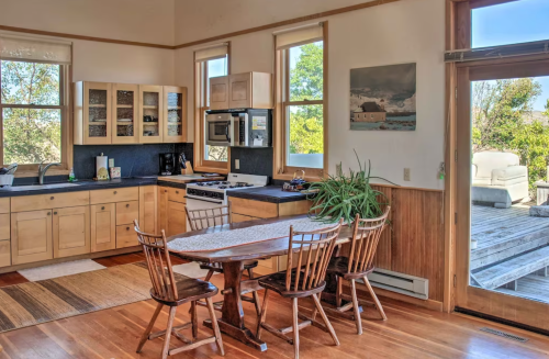 Bright kitchen with wooden cabinets, a dining table, and large windows overlooking a deck and greenery outside.