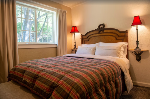 Cozy bedroom with a wooden bed, plaid comforter, and red lamps beside a window with natural light.