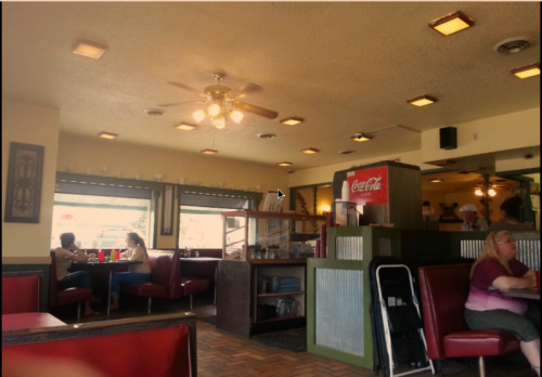 A cozy diner interior with red booths, a ceiling fan, and patrons enjoying their meals at tables.
