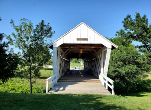 A white covered bridge surrounded by green trees and grass under a clear blue sky.