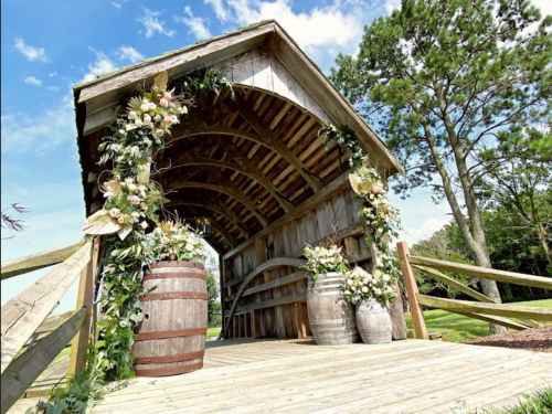 A rustic wooden gazebo adorned with flowers and greenery, flanked by two barrels, set against a blue sky.