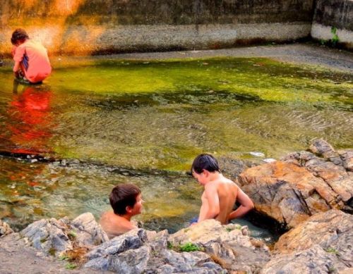 Three boys play in a natural pool surrounded by rocks, with one sitting on the edge and two splashing in the water.