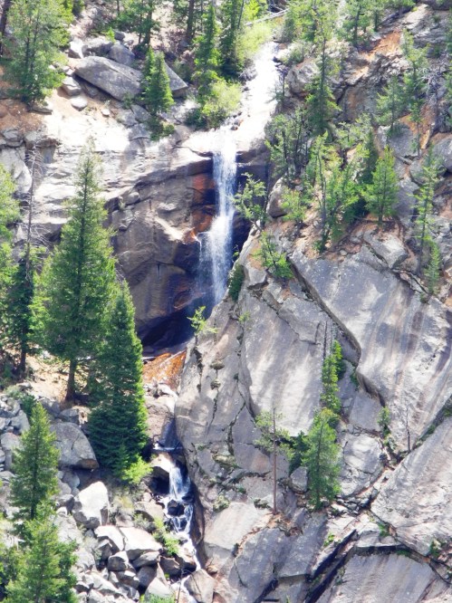 A waterfall cascades down rocky cliffs, surrounded by lush green trees in a mountainous landscape.