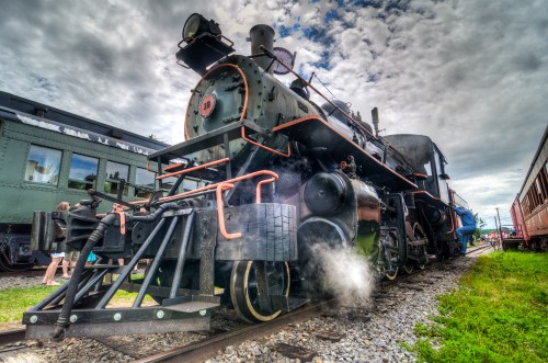 A vintage steam locomotive on tracks, with clouds overhead and people in the background, releasing a puff of steam.