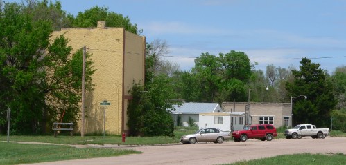 A small town scene featuring a yellow building, several parked cars, and green trees under a blue sky.