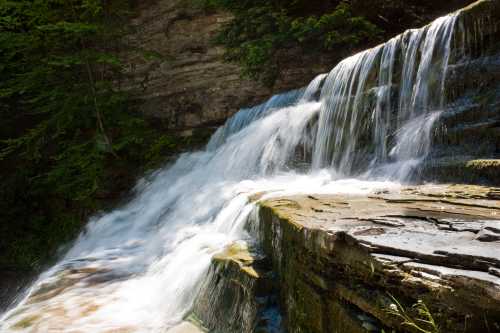 A serene waterfall cascading over rocky ledges, surrounded by lush greenery and sunlight filtering through the trees.