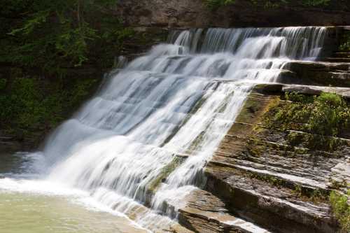 A serene waterfall cascading over rocky steps, surrounded by lush greenery and a calm pool below.