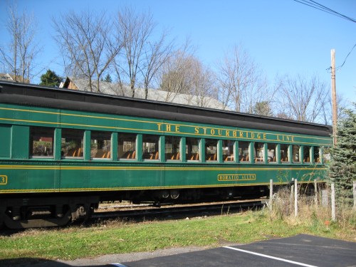 A green vintage train car labeled "The Stourbridge Line" parked near trees and a building on a sunny day.