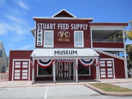 Red wooden building with "Stuart Feed Supply" sign, featuring a museum entrance and patriotic decorations.