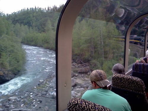 View from a train window showing a river and lush greenery, with passengers seated inside the train.