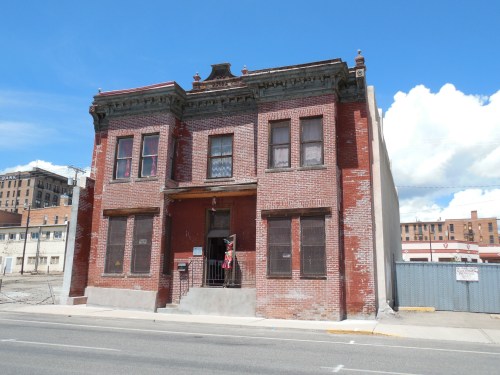 A two-story brick building with ornate details, featuring boarded windows and a central entrance, under a blue sky.