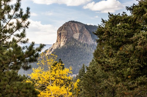 A rocky mountain peak rises above a forest of green trees and autumn-colored foliage under a cloudy sky.
