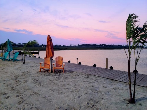 Two wooden chairs and an umbrella on a sandy beach at sunset, with calm water and palm trees in the background.