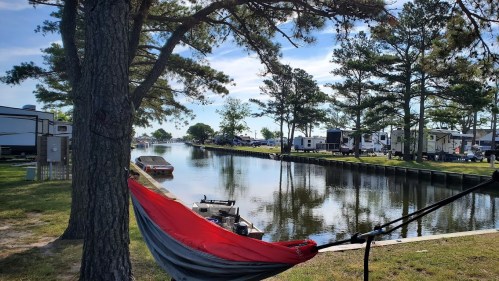 A peaceful canal scene with a hammock hanging between trees, boats docked, and RVs in the background under a clear sky.
