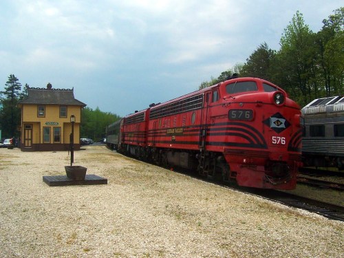 A vintage red train labeled "576" parked beside a small yellow train station in a scenic outdoor setting.