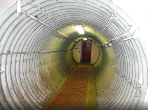 A long, cylindrical tunnel with metal walls and a grated floor, leading to a door at the far end.