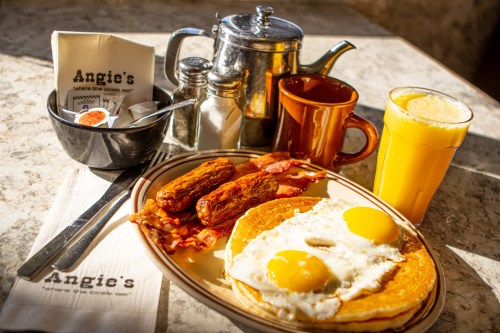 A hearty breakfast plate with eggs, bacon, sausage, orange juice, and a coffee pot on a table at Angie's.