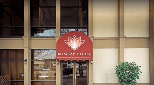 Exterior of Bombay House restaurant featuring a red awning with a lotus logo and large glass windows.