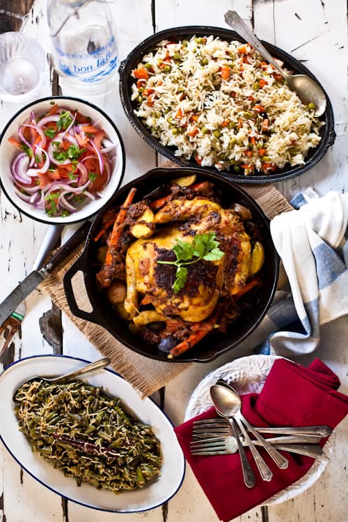 A table spread featuring roasted chicken, rice, salad, and greens, with utensils and a bottle of water.