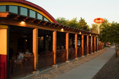 A colorful restaurant exterior with wooden pillars, outdoor seating, and a vintage sign at sunset.