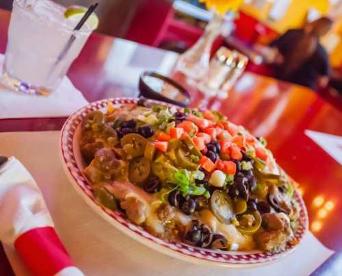 A colorful plate of nachos topped with jalapeños, olives, tomatoes, and green onions, served with a drink in the background.