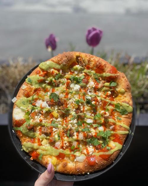 A hand holding a colorful pizza topped with avocado, cheese, herbs, and sauces, with flowers in the background.