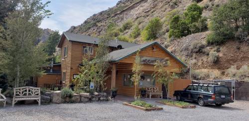 A rustic log cabin-style building surrounded by trees and mountains, with a gravel parking area and outdoor seating.