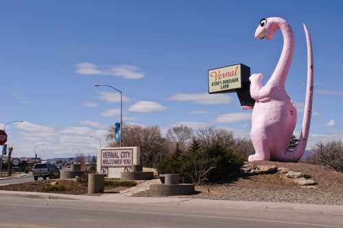 A large pink dinosaur statue stands beside a sign welcoming visitors to Vernal City, with blue skies in the background.