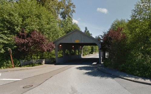 A covered bridge surrounded by trees, with a clear blue sky above and a road leading through it.