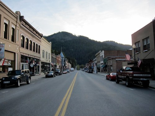 A quiet main street lined with historic buildings and parked cars, with mountains in the background under a clear sky.
