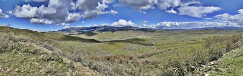 A panoramic view of rolling green hills under a blue sky with fluffy clouds and distant mountains.