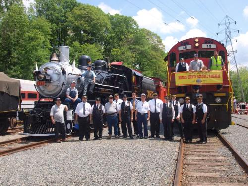 A group of people in uniforms poses with two vintage trains on a sunny day, surrounded by greenery.