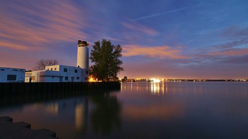 A serene lakeside scene at dusk, featuring a lighthouse, trees, and reflections on calm water.