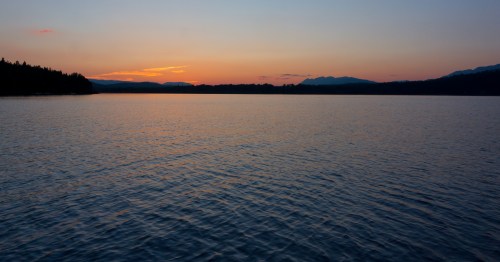 A serene lake at sunset, with calm waters reflecting the colorful sky and silhouettes of distant mountains.