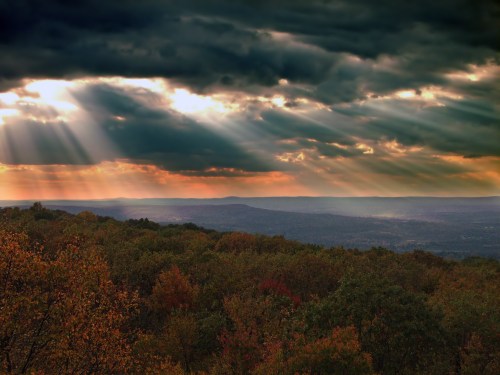 Dramatic sky with rays of sunlight breaking through clouds over a colorful autumn landscape.