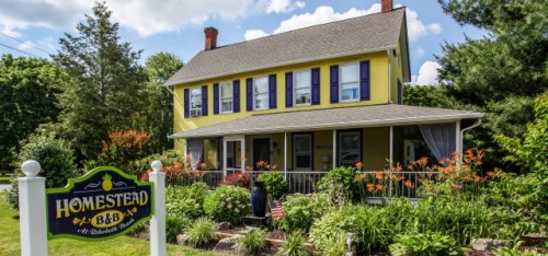 A charming yellow B&B with blue shutters, surrounded by lush greenery and flowers, featuring a welcoming sign.