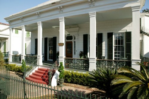 A historic white house with green shutters, a red staircase, and a wrought iron fence, surrounded by lush greenery.