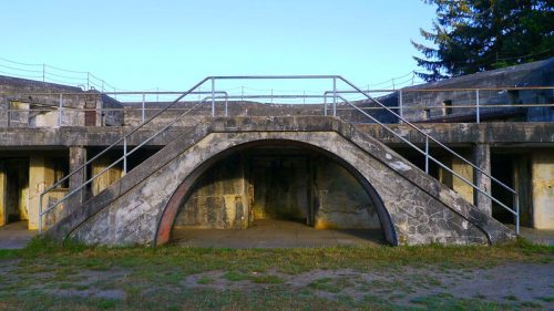 A weathered concrete structure with an arched entrance, surrounded by grass and trees under a clear blue sky.
