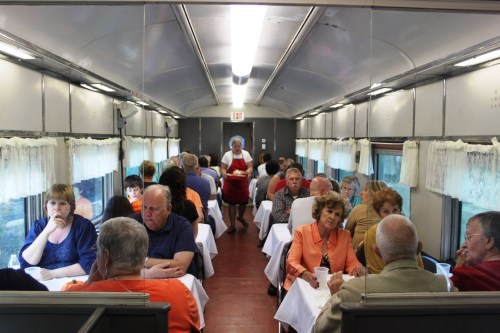 A dining car filled with people enjoying meals at tables, with a server walking through the aisle.