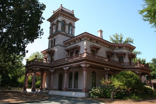 A historic Victorian-style mansion with a tower, surrounded by trees and greenery on a sunny day.