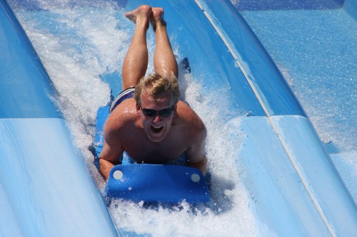 A man in sunglasses slides down a water slide, splashing into the water below with excitement.