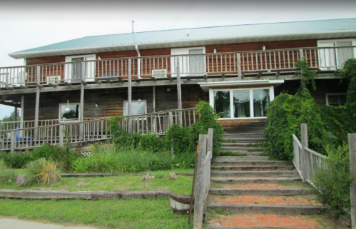 A two-story wooden building with a balcony, surrounded by greenery and a staircase leading to the entrance.
