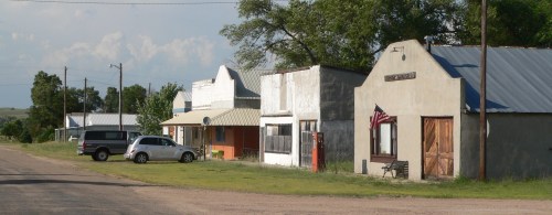 A quiet rural street lined with small, weathered buildings and a parked car, featuring an American flag on one storefront.